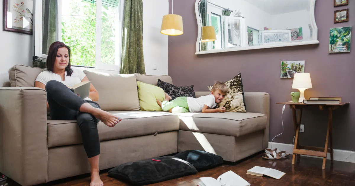 Woman reading on beige sectional sofa with child in cozy living room makeover featuring layered lighting and green accents